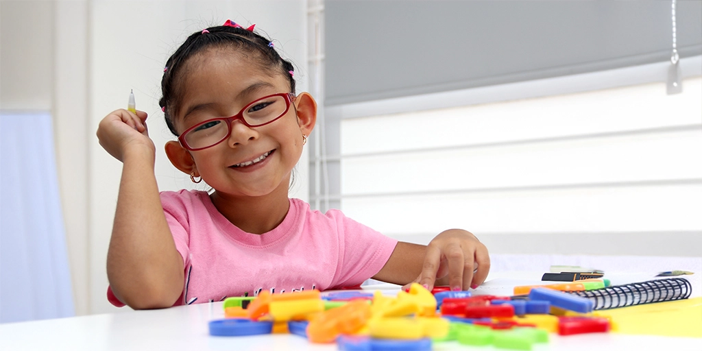 A young girl with glasses smiling brightly while working on colorful educational toys, representing creativity and positivity in supportive environments for children with autism.