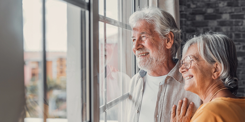 An elderly couple smiles while looking out of a large window in a cozy, modern living room, symbolizing comfort and security with the use of AngelSense Assistive Technology. 