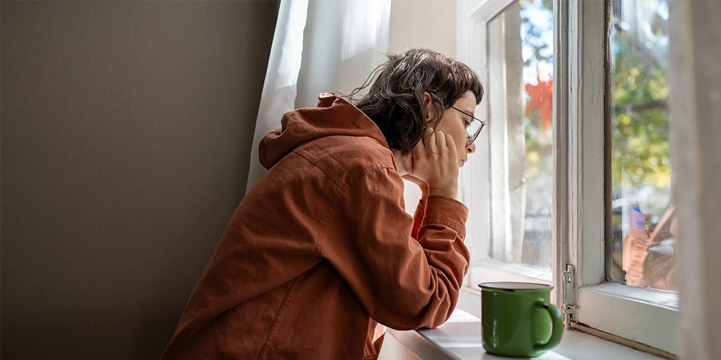 A person with autism wearing glasses and an orange hoodie, sitting by a window with a green mug beside them, looking thoughtfully outside.