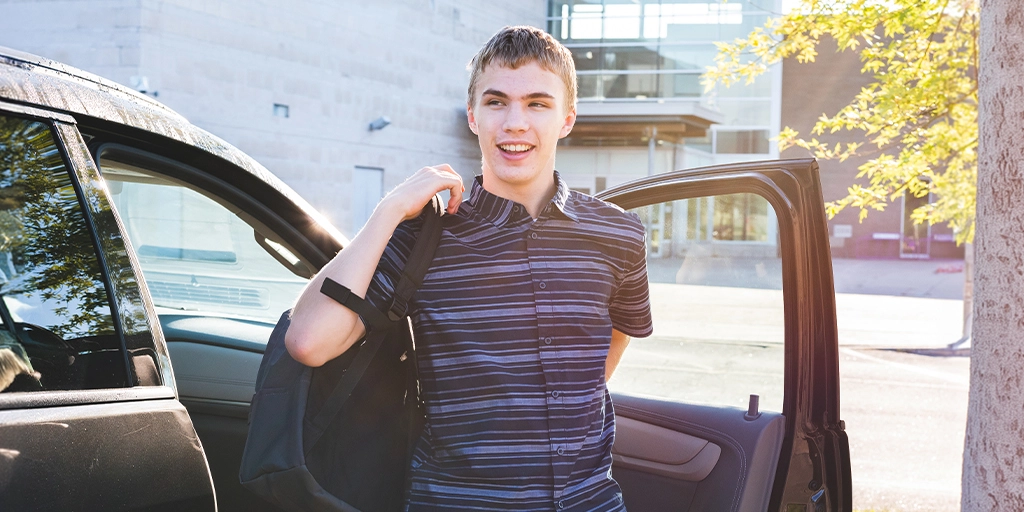 A young person with autism is standing beside an open car door, wearing a striped shirt and carrying a backpack, showing a moment of independence despite the autism challenges of worry and dependence.