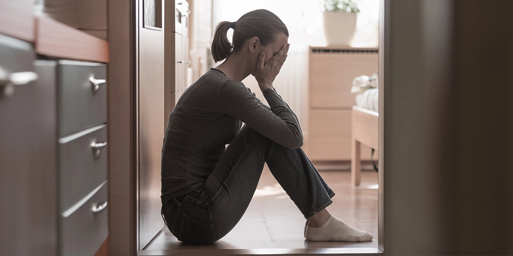 A mother sitting on the floor in a hallway, leaning against the wall with their head in their hands, depicts the stress and emotional toll of dealing with the vicious cycle of autism safety concerns.